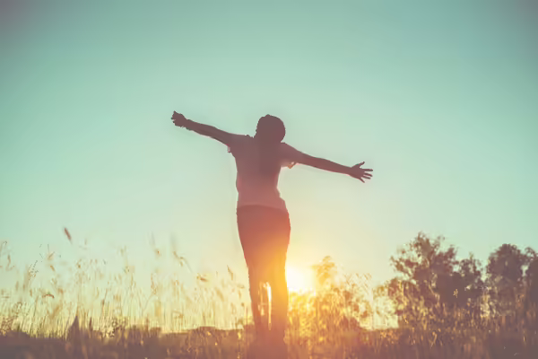 woman standing in a field with arms wide open and sun setting 