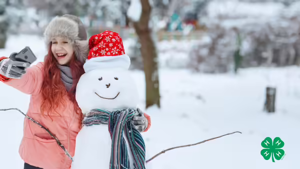 A smiling girl taking a selfie with a snowman. A green 4-H logo in the lower right corner.