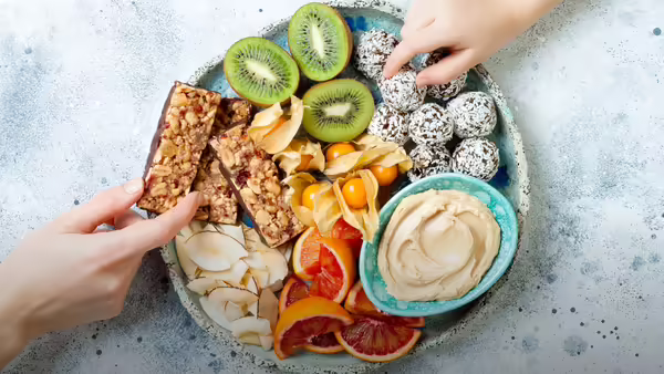 Overhead view of two hands sharing a platter of healthy snacks, including fruit, energy balls, and dips.