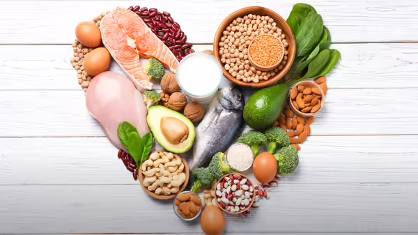 A heart shape made of foods including fish, chicken, eggs, nuts, beans, lentils, avocado, broccoli, and spinach.