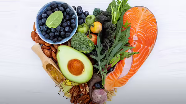 A heart-shaped arrangement of heart-healthy foods, including salmon, blueberries, almonds, avocado, broccoli, kale, and other nuts and vegetables.