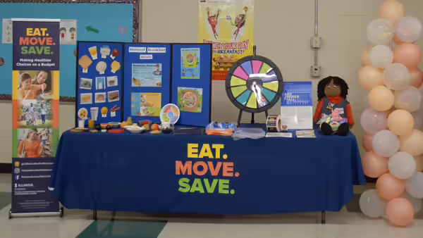 A table covered with a blue tablecloth featuring the Eat. Move. Save. logo, displaying games and informational flyers, with balloons positioned nearby.