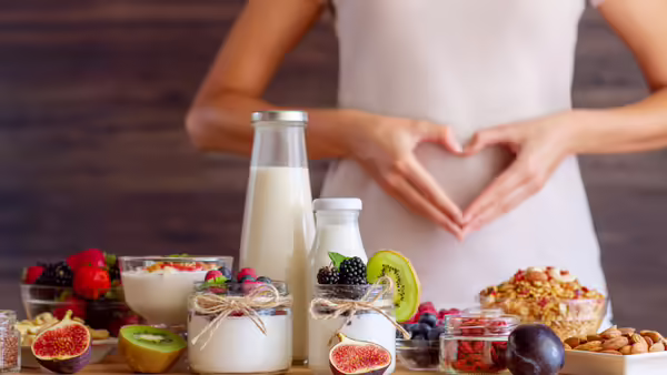A table with yogurt jars topped with fresh fruits, nuts, and seeds, and there is a person who is forming a heart shape with their hands over their stomach.
