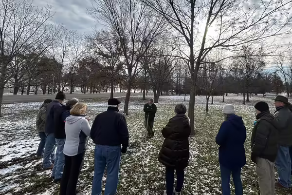 A man leans on a tool outside by a tree surrounded by a group of people