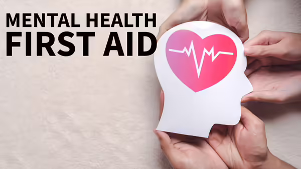 Text reads "Mental Health First Aid". On the right, several hands hold a white paper cutout of a human head silhouette featuring a pink heart.