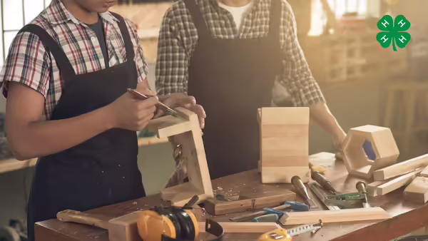 An adult and a child wearing aprons work together at a woodworking bench filled with tools. The 4-H logo is in the top right corner.