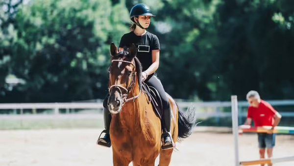 girl riding a horse in an arena, wearing a helmet, with someone in the background moving a fence