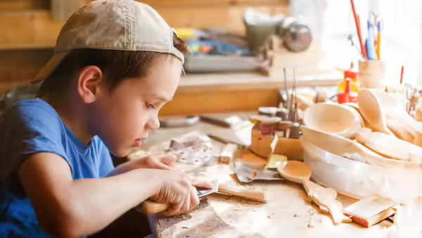 A young boy wearing a backwards baseball hat witling wood.