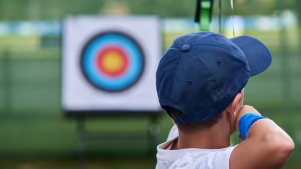 A little kids with a navy blue hat aiming at an archery target.