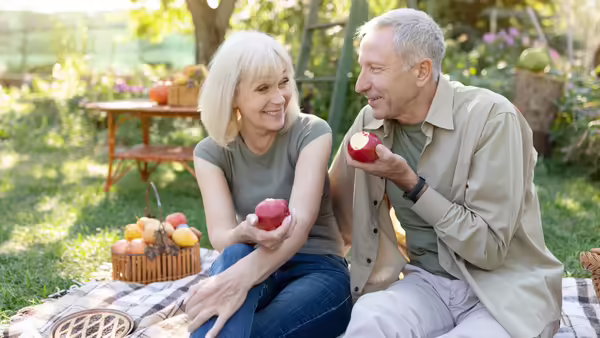 An older couple eating red apples while they sit on a picnic blanket.
