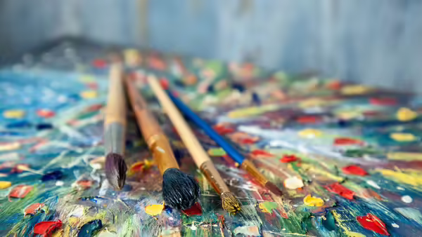 Various sized paintbrushes laying on a rainbow paint splattered table.