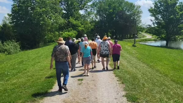 Group of adults walking together along a gravel path in a sunny park, with green grass, trees, and a pond beside a winding road in the background.
