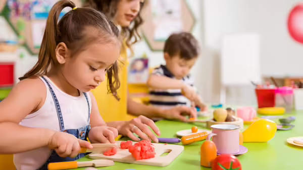 Children at a play table using toy food and utensils to pretend-cut vegetables during a kitchen-themed activity.
