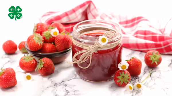 A jar of strawberry jam with strawberries on the table. A green 4-H logo in upper left corner.