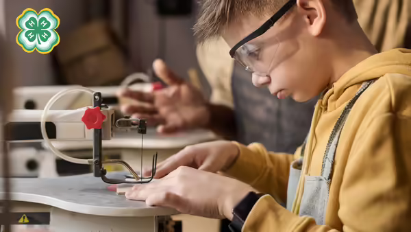 A boy with safety glasses guiding a piece of wood through a saw. A green 4-H clover in the upper left corner.