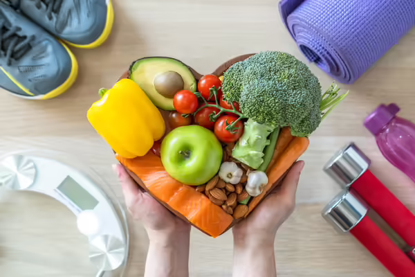 Heart shaped bowl with fruits and veggies surrounded by workout equipment