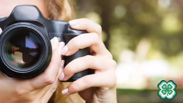Close-up of a person holding a camera to their eye, with a green 4-H clover logo in the bottom right corner and a blurred outdoor background.