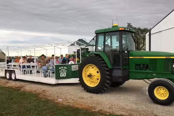 People riding on a seated trailer being pulled around a field day event by a green tractor.