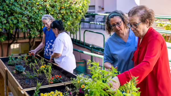 An elderly woman and a healthcare worker in scrubs tending to a raised garden bed together outdoors