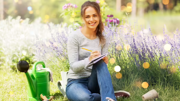 A woman sitting on the grass in a garden, writing in a notebook next to a green watering can and purple flowers.