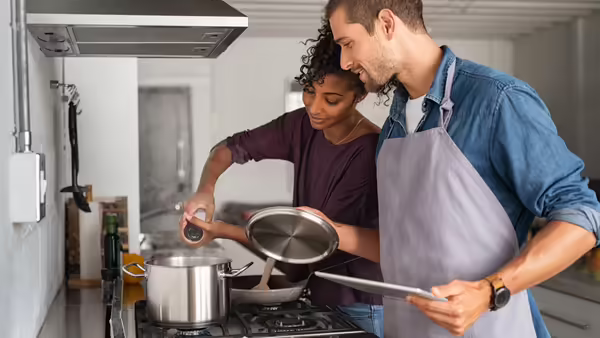 Woman adding salt in pot while cooking while a man looks over her shoulder holding a pan lid.