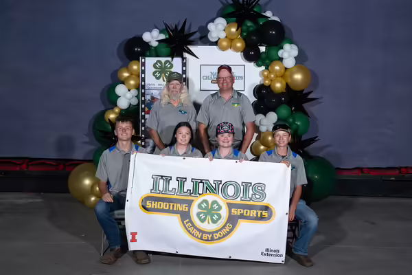 two standing adults and four seated youth holding a banner that says "Illinois Shooting Sports, Learn By Doing"