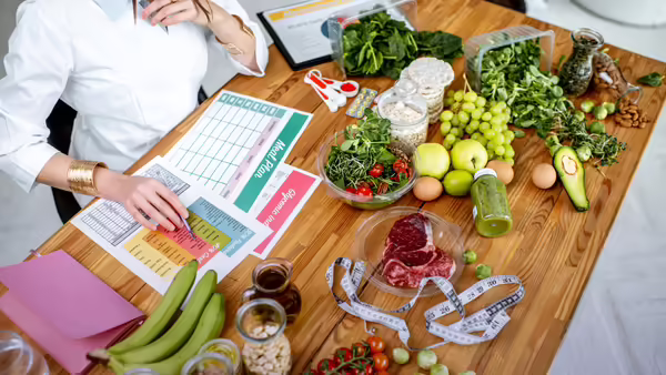A person sitting at a wooden desk filled with fresh healthy food, measuring tape, and meal-planning charts.