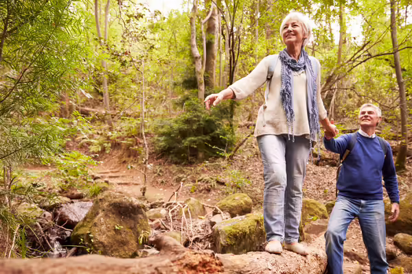 In the woods, a white-haired woman balances on a log while holding a man's hand.