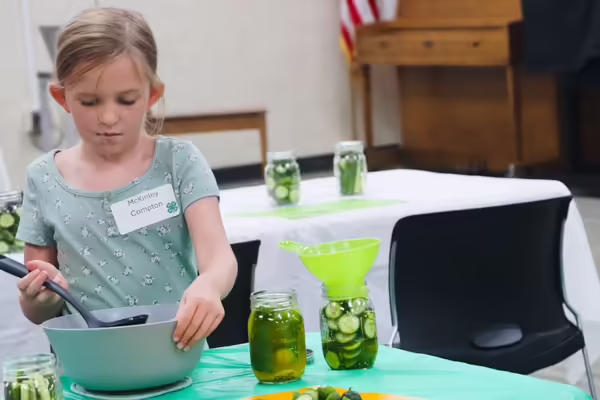 A young girl uses a ladle to pour pickle brine into a jar of cucumbers.