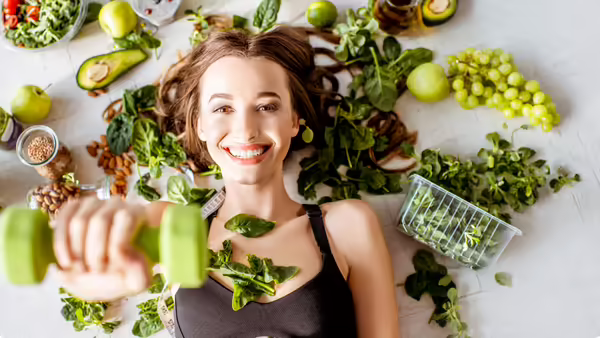 A smiling woman in workout clothing holds a green dumbbell while surrounded by fresh fruits, vegetables, leafy greens, and other healthy foods.