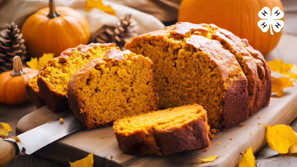 Sliced loaf of pumpkin bread on a cutting board, surrounded by whole pumpkins, pinecones, and autumn leaves, with a 4-H logo in the top right corner.