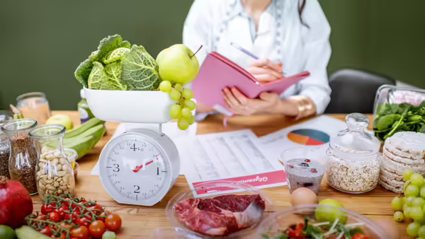 A person writing in a notebook behind a table arranged with fresh produce, raw meat, grains, and a food scale.