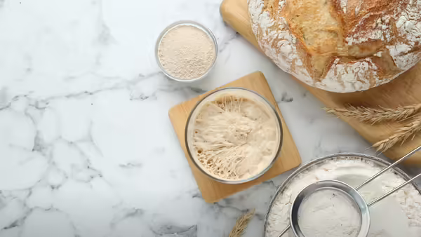 A white marble surface, including a jar of active starter, a bowl of flour with a sifter, a bowl of yeast, wheat stalks, and a freshly baked sourdough loaf.