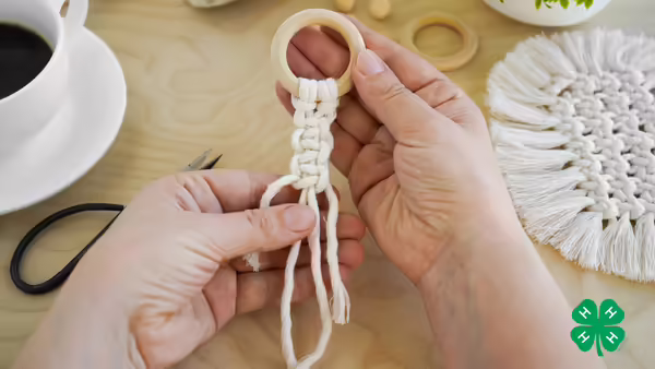 Hands crafting a macramé design attached to a wooden ring on a wooden tabletop next to a coffee cup and scissors. Green 4-H clover in bottom right corner.