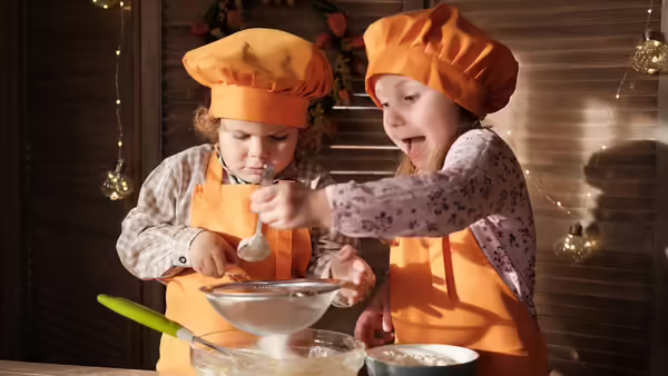 two young kids cooking and holding a spoon and sifter while wearing orange aprons and cooking hats