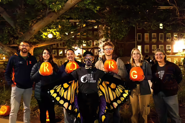 Seven people at night in front of a college residence hall. Four are holding pumpkins. One is dressed like a butterfly.