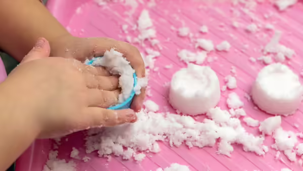 a close-up of a kids hands packing snow in a container with snow nearby on a pink mat