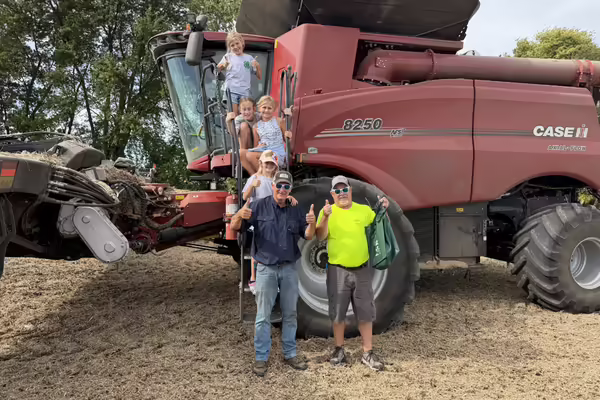 Youth sit on a combine while two farmers wearing sunglasses give thumbs up signs to the camera
