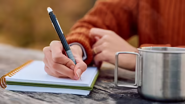 A person writing in a notebook next to a metal mug on a wooden table.
