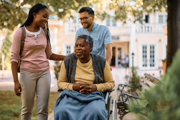 older adult getting pushed in wheelchair by aid while talking with a family member 
