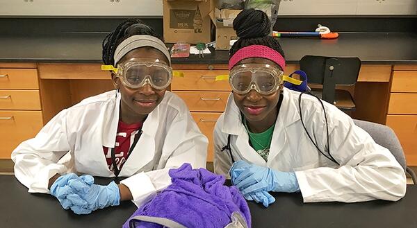 two girls wearing lab coats and safety goggles