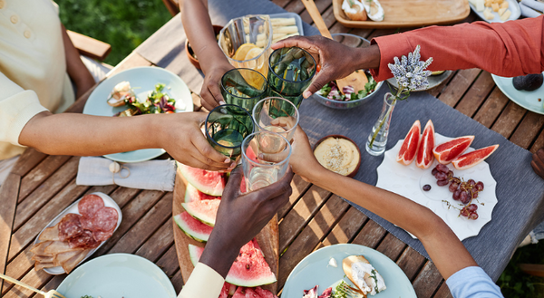 Hands on an African American family raising glasses in a toast over a table of summer foods