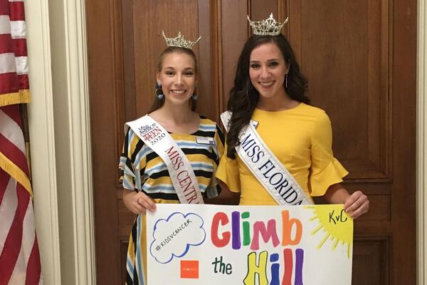 Two young women wearing crowns and sashes hold a sign that says "Climb the Hill" while lobbying. 