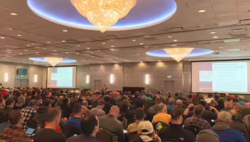 A large room completely filled with people sitting in chairs at tables listening to a training presentation.