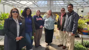 Six people, some wearing branded University of Illinois apparel, stand inside a greenhouse