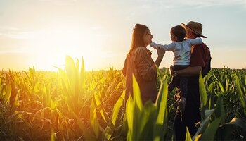 A woman stands next to her husband holding their toddler in the middle of a corn field. The sun is setting in the distance, casting a golden hue, and a serene image. 