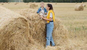 A woman standing next to a child that is sitting on top of a bail of hay