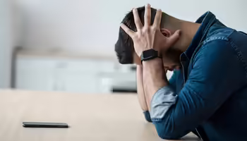 Man in a denim shirt sits, elbows on a table with head rested in his hands.