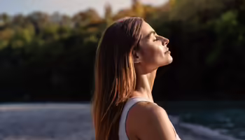 Women standing by water meditating with sun shining on her face with trees in the background