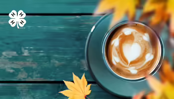 A cup of a latte on a teal wooden table with orange and yellows leaves around the cup. In the top left corner is a white 4-H logo.